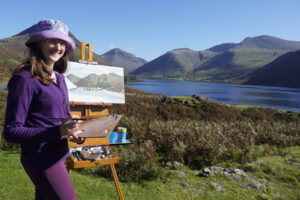 Artist katie stood outside holding painting pallet and brush in front of easel with half painted canvas on. In the background is lake and mountains