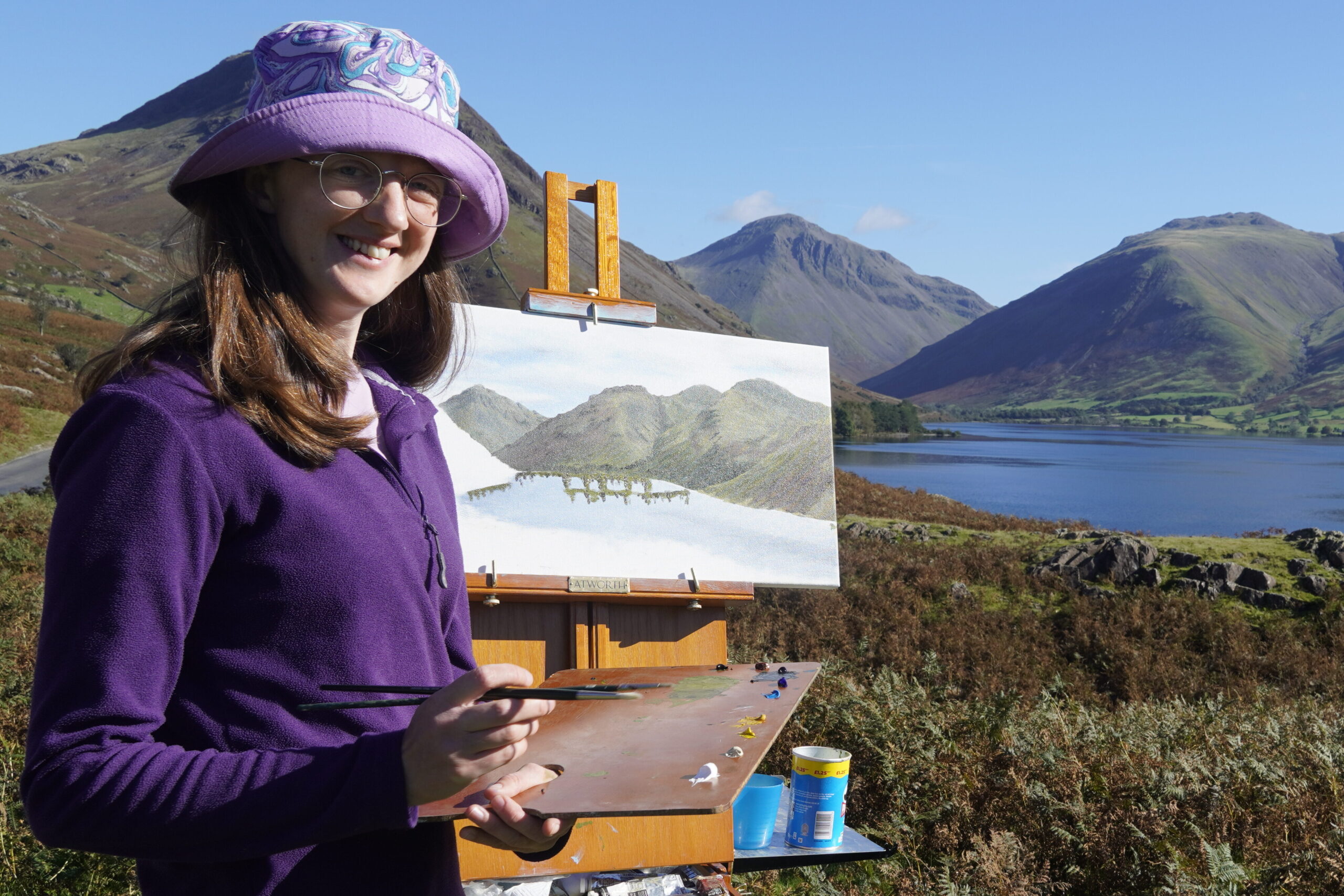 Image of the artist stood holding a painting pallet with a half finished painting on an esale and the landscape of mountians and lake in the background